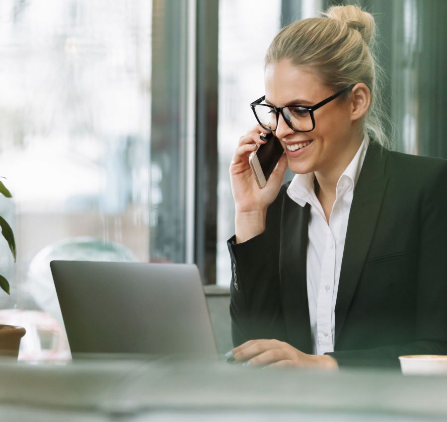 Photo of young happy smiling blonde business woman sitting indoors in cafe using laptop computer talking by mobile phone. Looking aside.