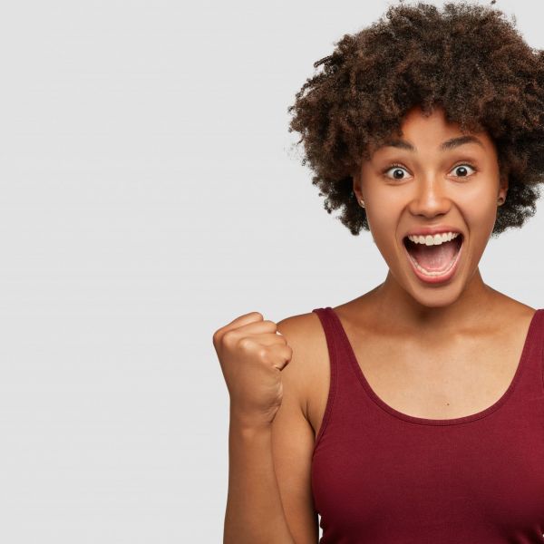 Dark skinned triumphing girl raises fists happily, celebrates victory, dressed in casual clothes, isolated over white background with free space for your promotion. Student rejoices finish of semester