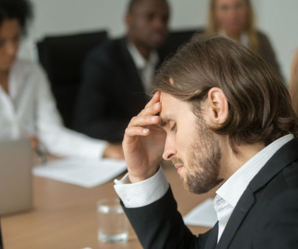 Frustrated tired businessman having headache at team meeting, upset company ceo feeling unwell suffering from migraine touching forehead at work, aching head or chronic fatigue in office concept