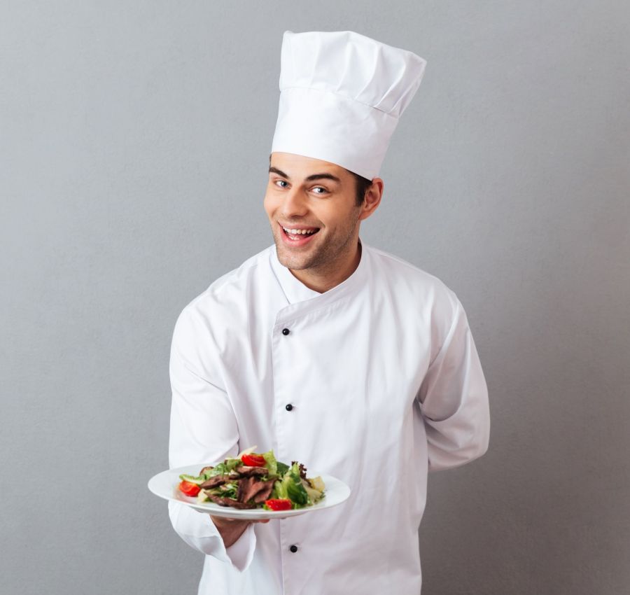 Picture of happy young cook in uniform standing isolated over grey wall background. Looking camera holding salad.